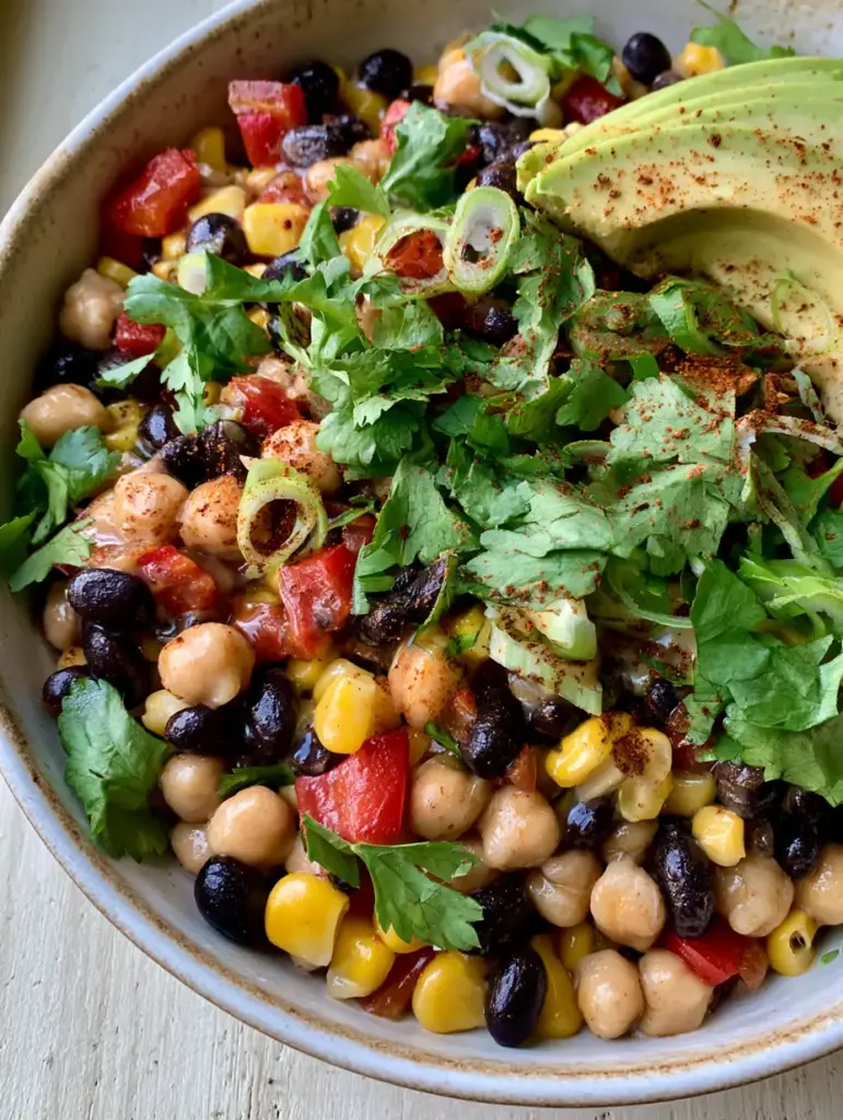 Southwest Chickpea Black Bean Salad with Zesty Lime Cumin Dressing 1 Rustic white bowl filled with Southwest Chickpea Black Bean Salad garnished with cilantro and avocado slices.