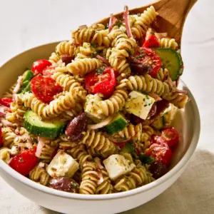 High angle shot of wooden servers lifting cold pasta salad from a white bowl.