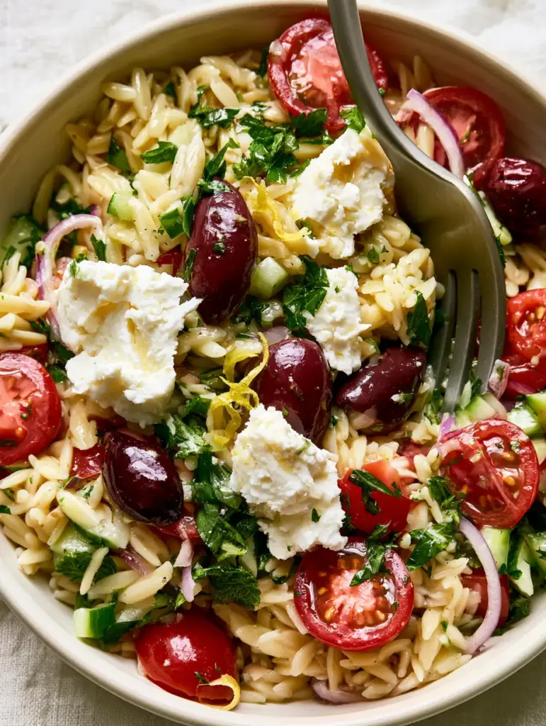 Greek Orzo Pasta Salad being tossed with a matte silver fork, highlighting the firm, non-clumping pasta texture.